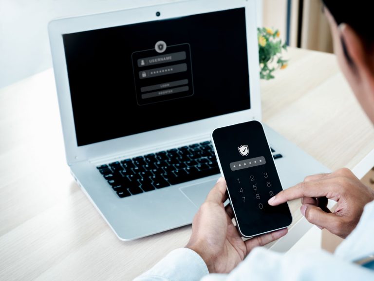Person typing passcode into a mobile phone app, with laptop in background displaying login screen