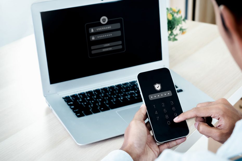 Person typing passcode into a mobile phone app, with laptop in background displaying login screen